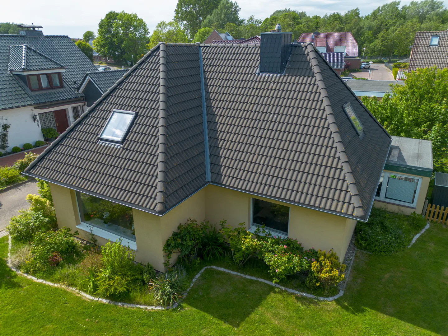 Aerial view of a beige house with a dark tiled roof, skylights, and a lush green garden in a suburban neighborhood.
