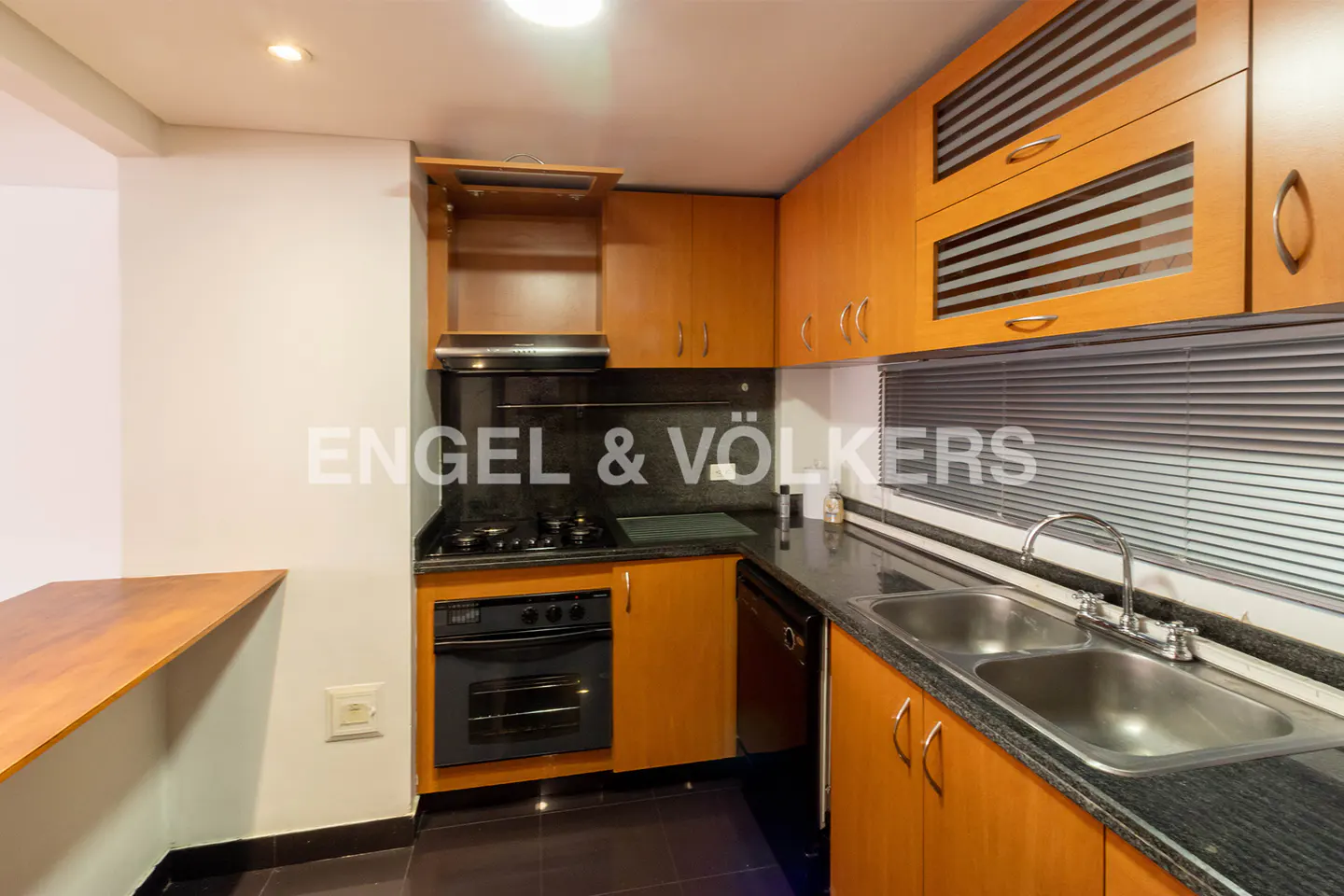 A kitchen with wood cabinets, black countertops, and stainless steel sink. A black oven is built into the cabinets.