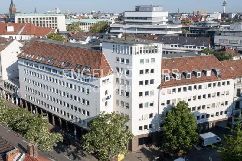 Aerial view of a white, multi-story building with a red-tiled roof in an urban setting. Trees and other buildings are visible in the background.