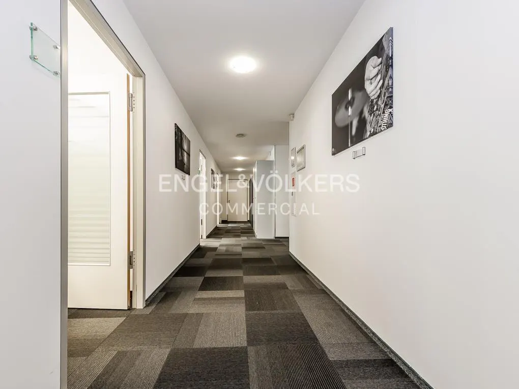 A long hallway with white walls and a gray patterned carpet. Black and white photos hang on the walls. Doors are visible along the hallway.