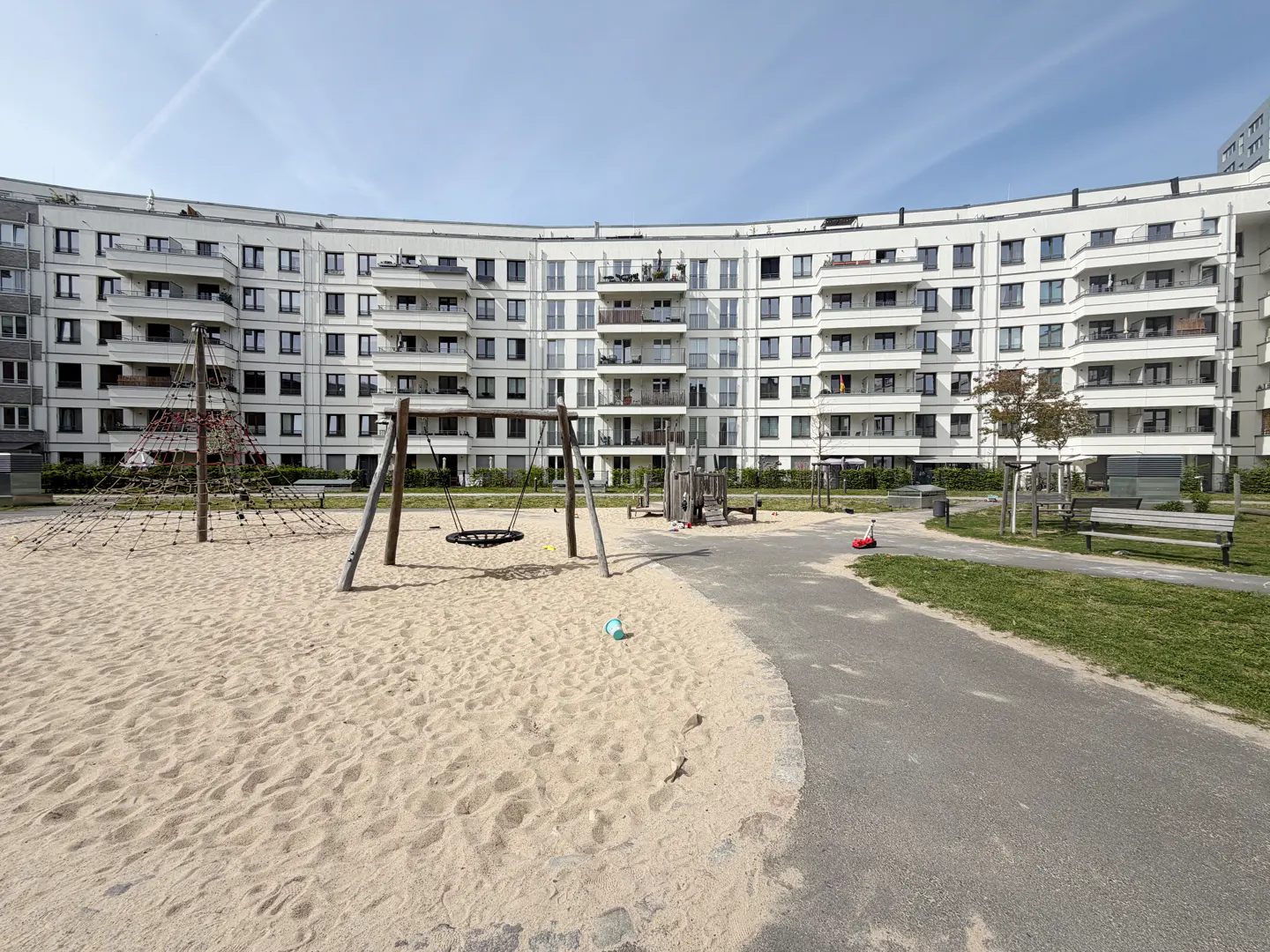 A curved, white apartment building overlooks a sandy playground with swings and climbing structures. A paved path leads to benches and green space.