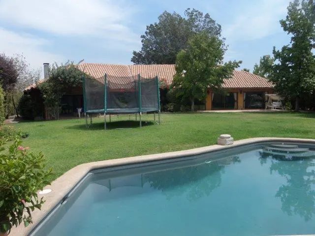 A backyard with a pool, green lawn, trampoline, and a house with a terracotta roof under a blue sky.