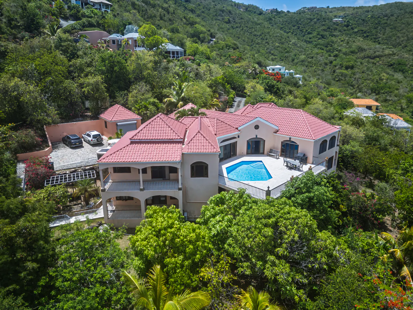 Aerial view of a beige villa with a red tile roof, private pool, and patio, nestled in a lush, green tropical hillside.
