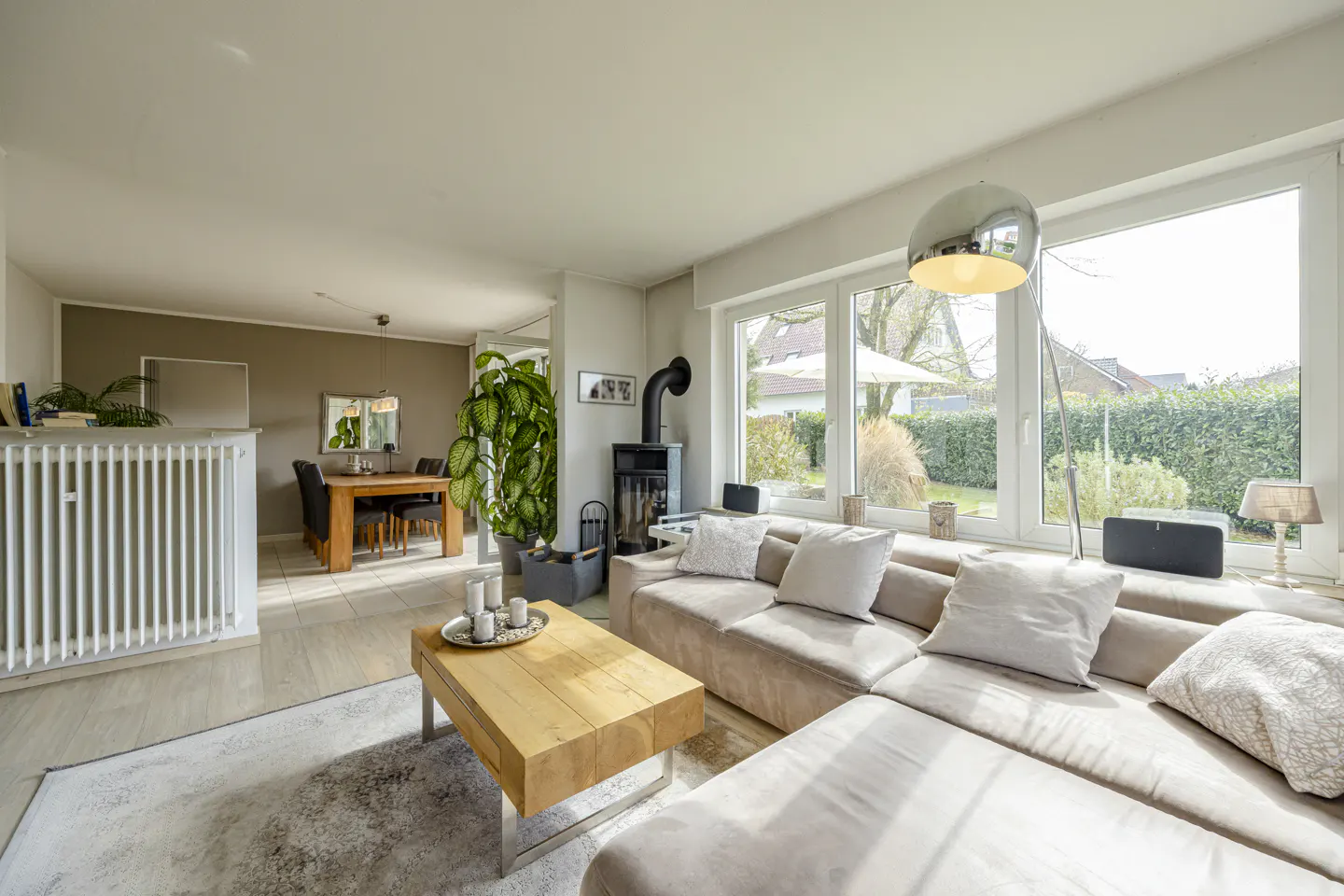 Living room with beige sectional sofa, wood coffee table, and black fireplace. Large windows overlook a green backyard. Dining room visible in background.