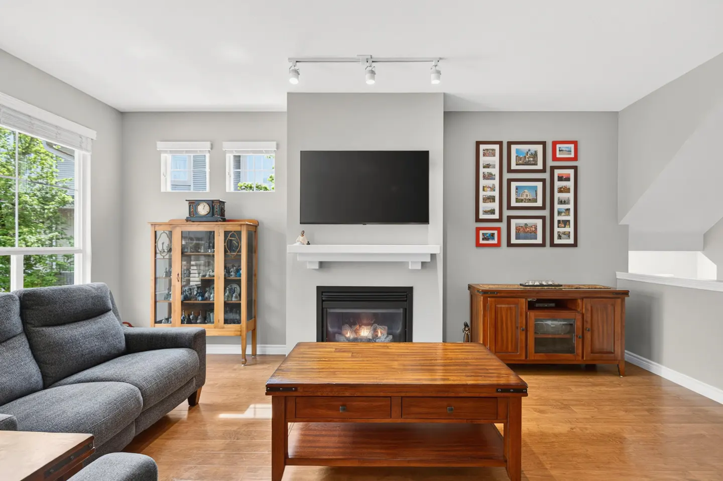Living room with gray sofa, wood floors, and a fireplace with a TV above it. A wooden coffee table sits in front of the fireplace.