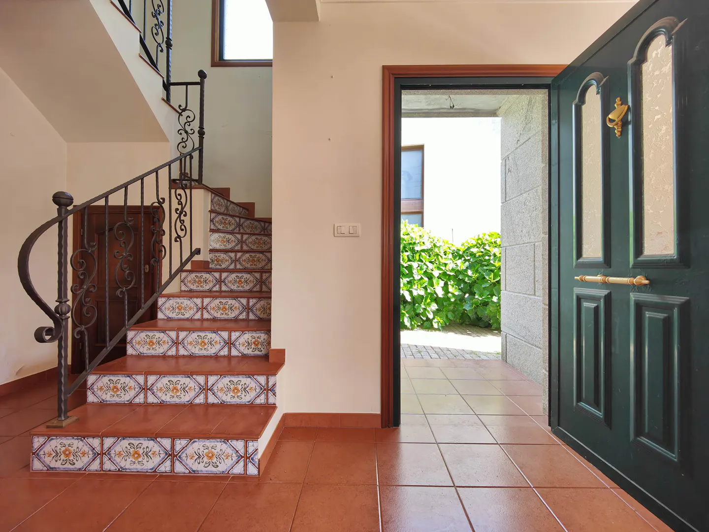 Foyer view with terracotta tile floor, stairs with floral tile risers, and an open dark green front door leading to a garden.