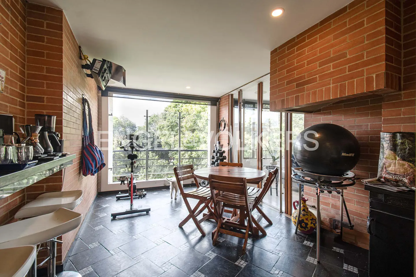 Brick-walled room with exercise bike, table, chairs, and exercise ball. Large windows overlook green trees.