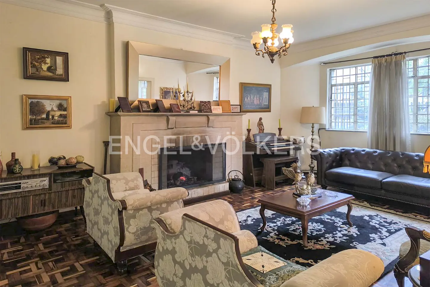 Living room with fireplace, floral armchairs, and a black leather sofa. A chandelier hangs from the ceiling.