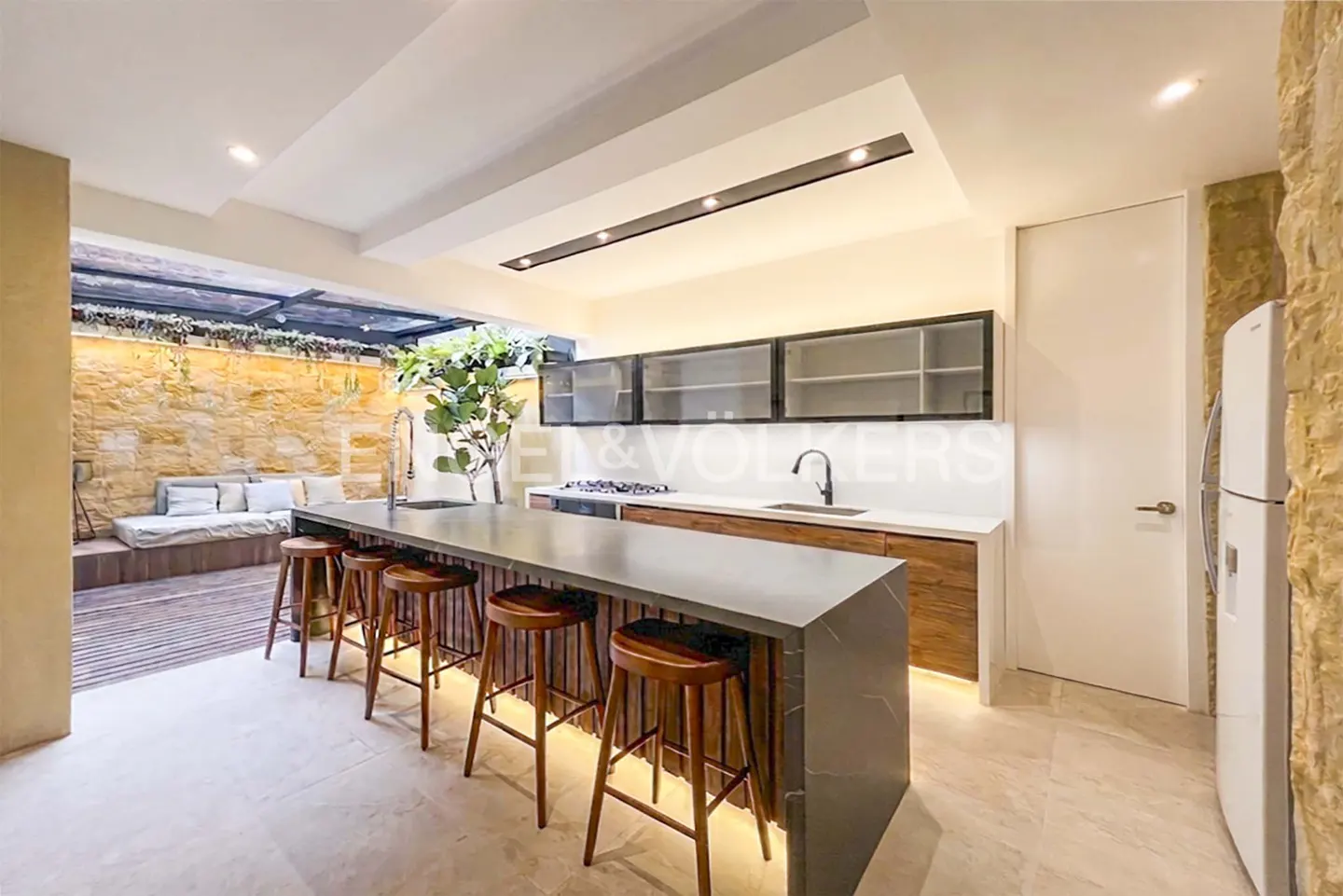 A modern kitchen with a long island, wooden stools, and an outdoor seating area with a stone wall.