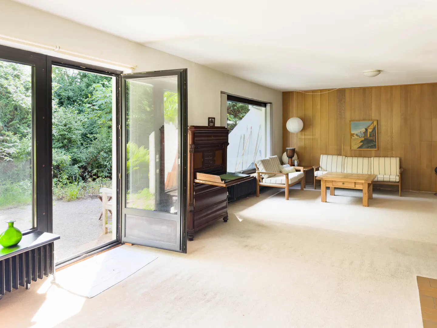 Bright living room with wood-paneled walls, a sofa, coffee table, antique cabinet, and glass doors opening to a garden.