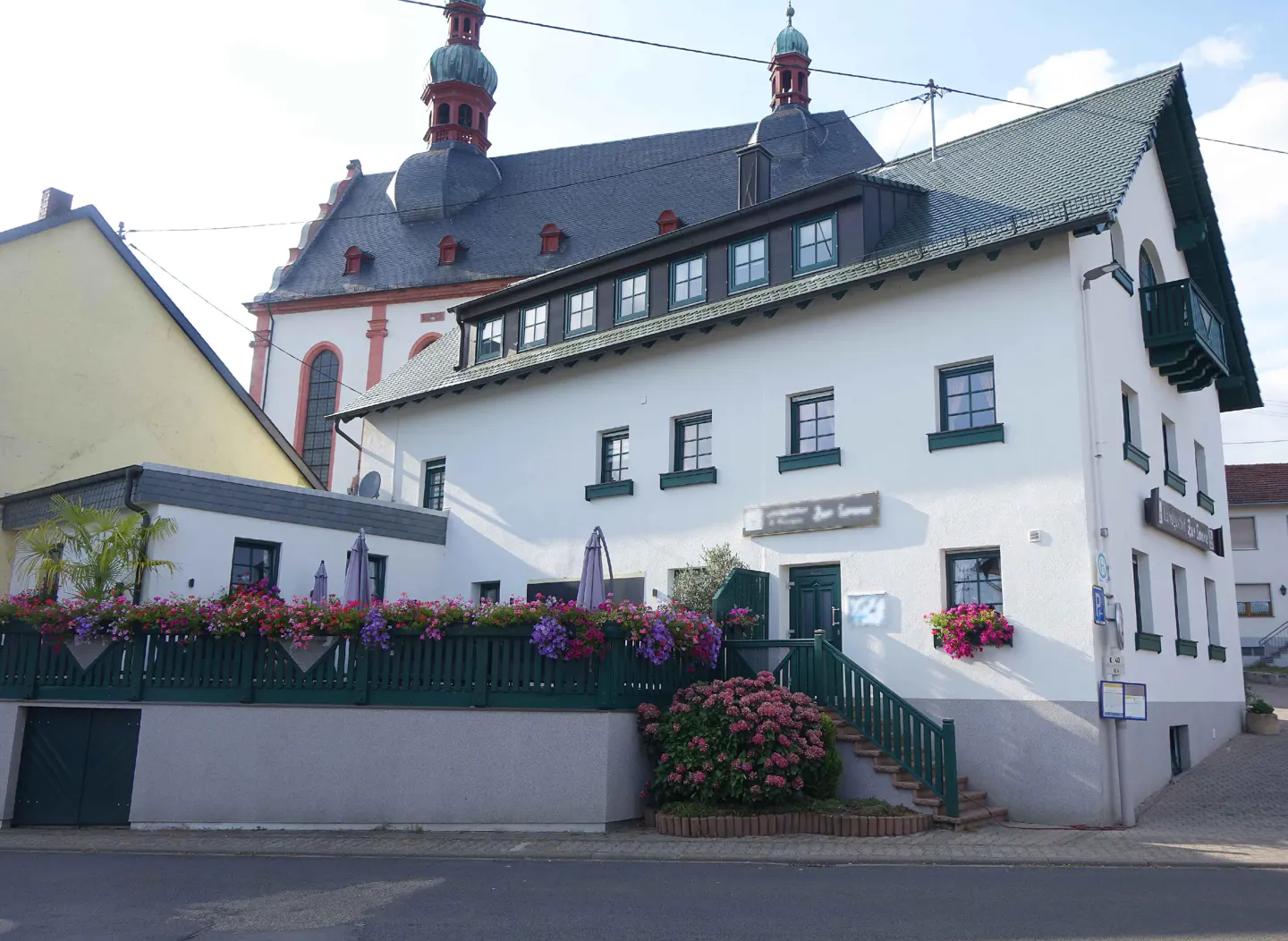 A white two-story building with a green roof and a church in the background.