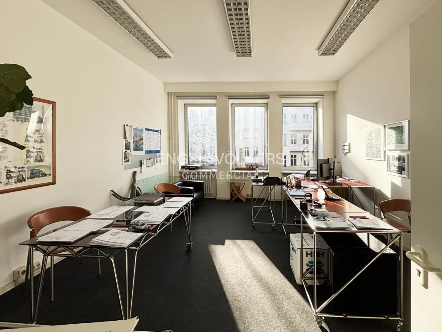 Bright office space with black floor, white walls, and large windows. Desks with papers, chairs, and a black couch furnish the room.