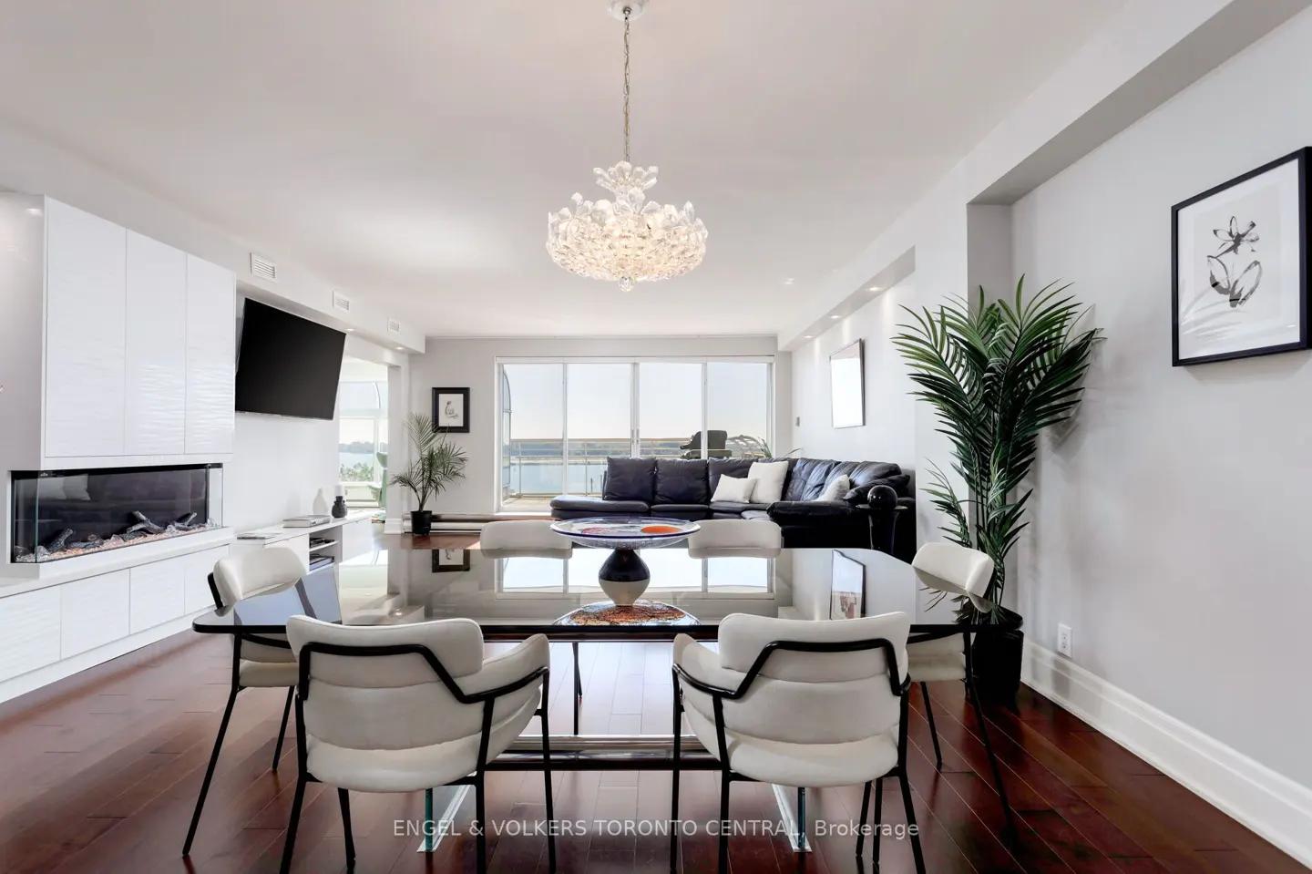 Bright, modern condo interior with a glass dining table, white chairs, and a crystal chandelier. A black leather sofa sits by large windows.