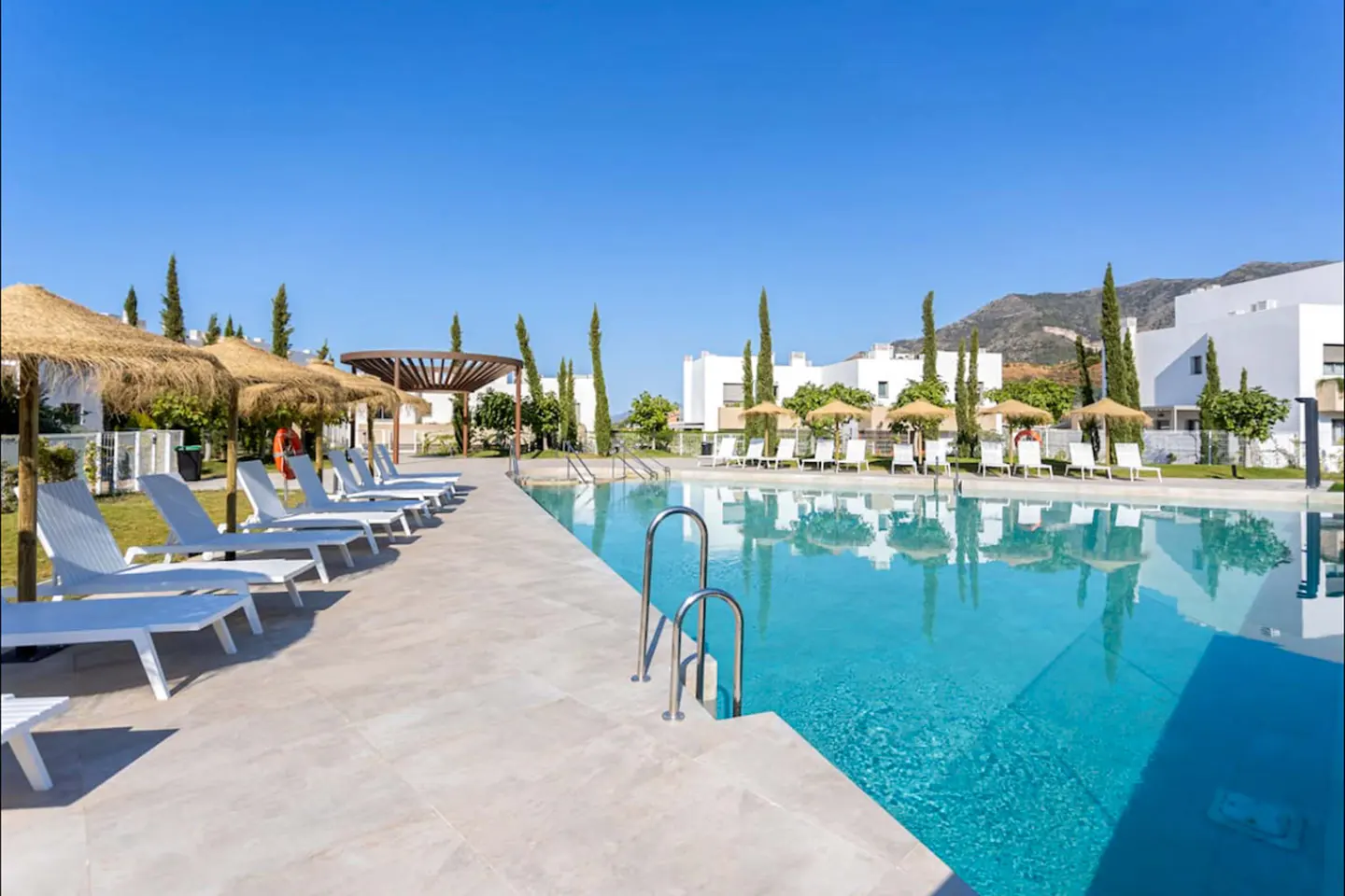 A bright, sunny view of a pool with white lounge chairs and straw umbrellas. White buildings and trees are in the background.