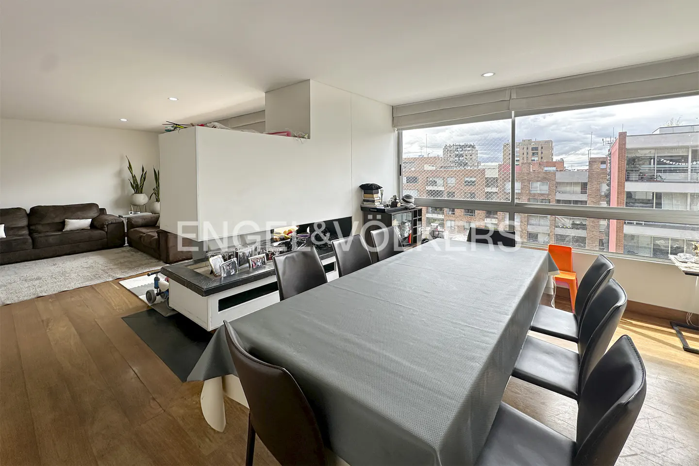 Open-concept living and dining area with wood floors, brown sofas, and a large gray dining table. City view through large windows.