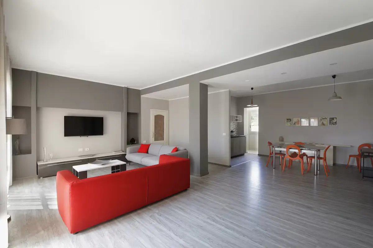 Open-concept living space with gray walls, wood floors, and a red sofa. A dining table with orange chairs is visible in the background.