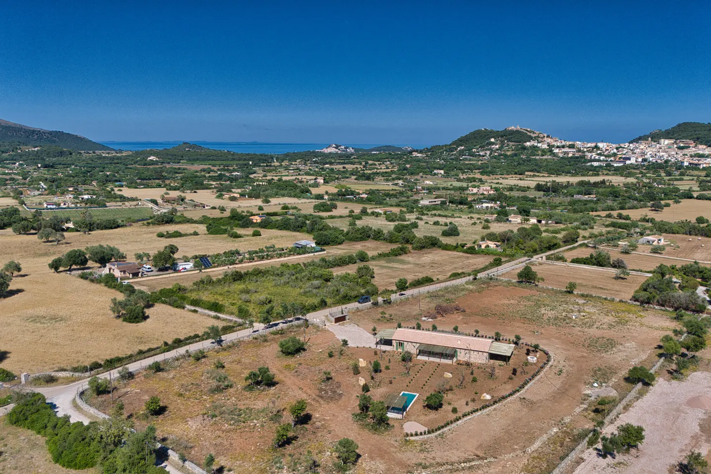 Aerial view of a tan, one-story house with a pool, surrounded by fields and trees under a blue sky.