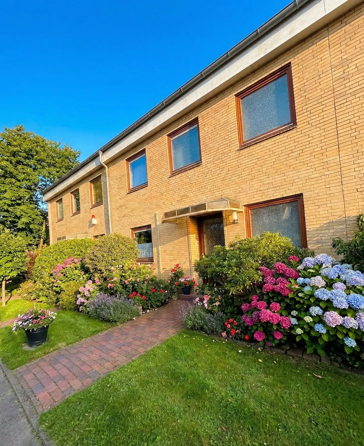 A two-story tan brick house with brown framed windows and a brick walkway lined with colorful flowers.
