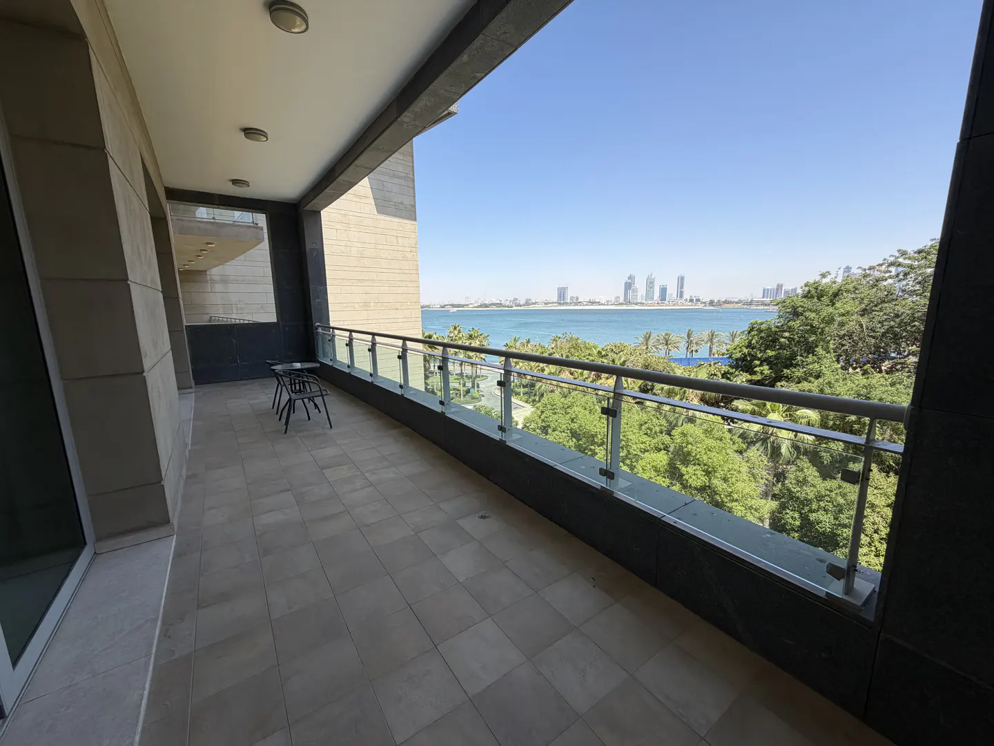 Spacious balcony with tiled floor, glass railing, and a scenic view of the ocean, lush trees, and a distant city skyline.