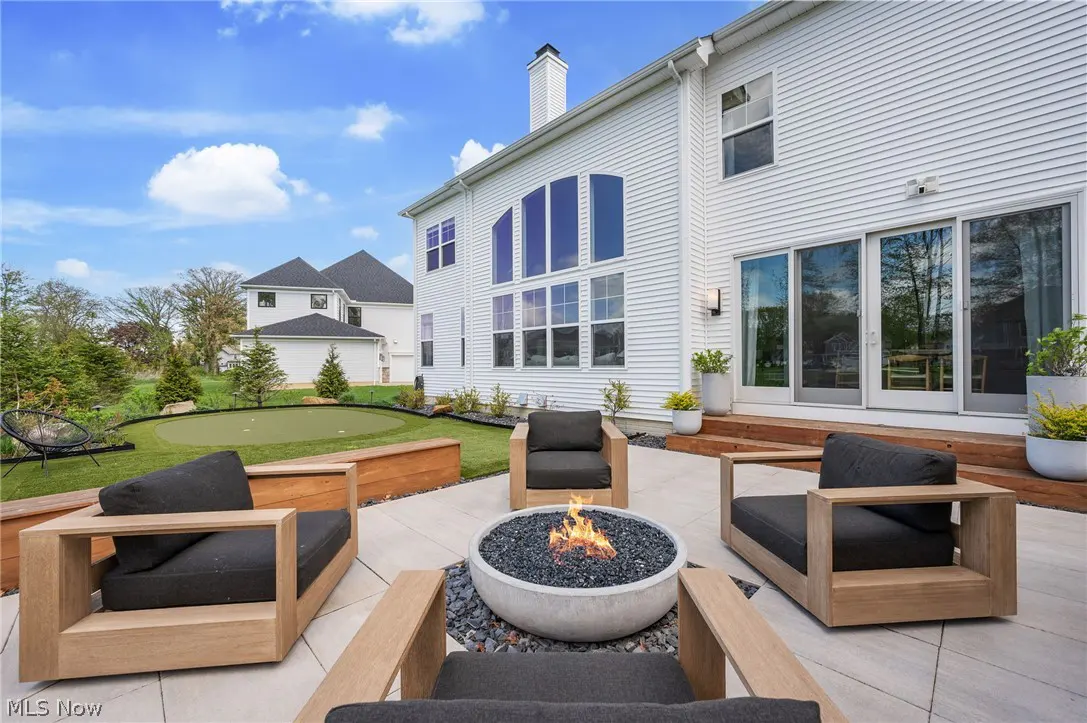 Outdoor patio with a fire pit surrounded by four chairs, a putting green, and a white house with large windows.