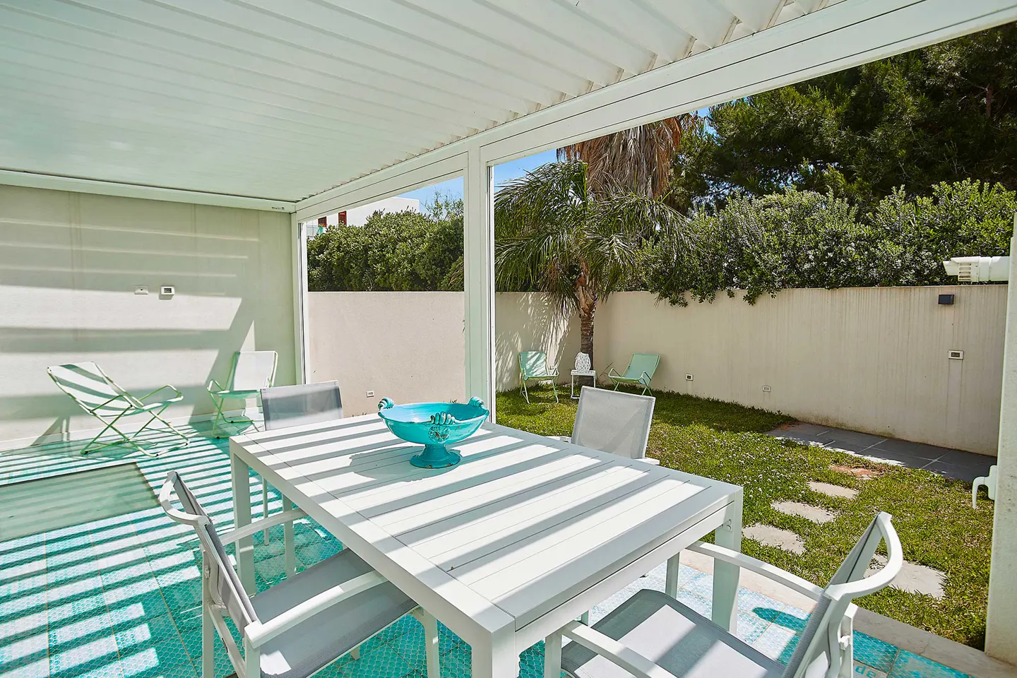 Covered patio with white dining set, turquoise bowl, and chairs overlooking a sunny backyard with green lawn and trees.