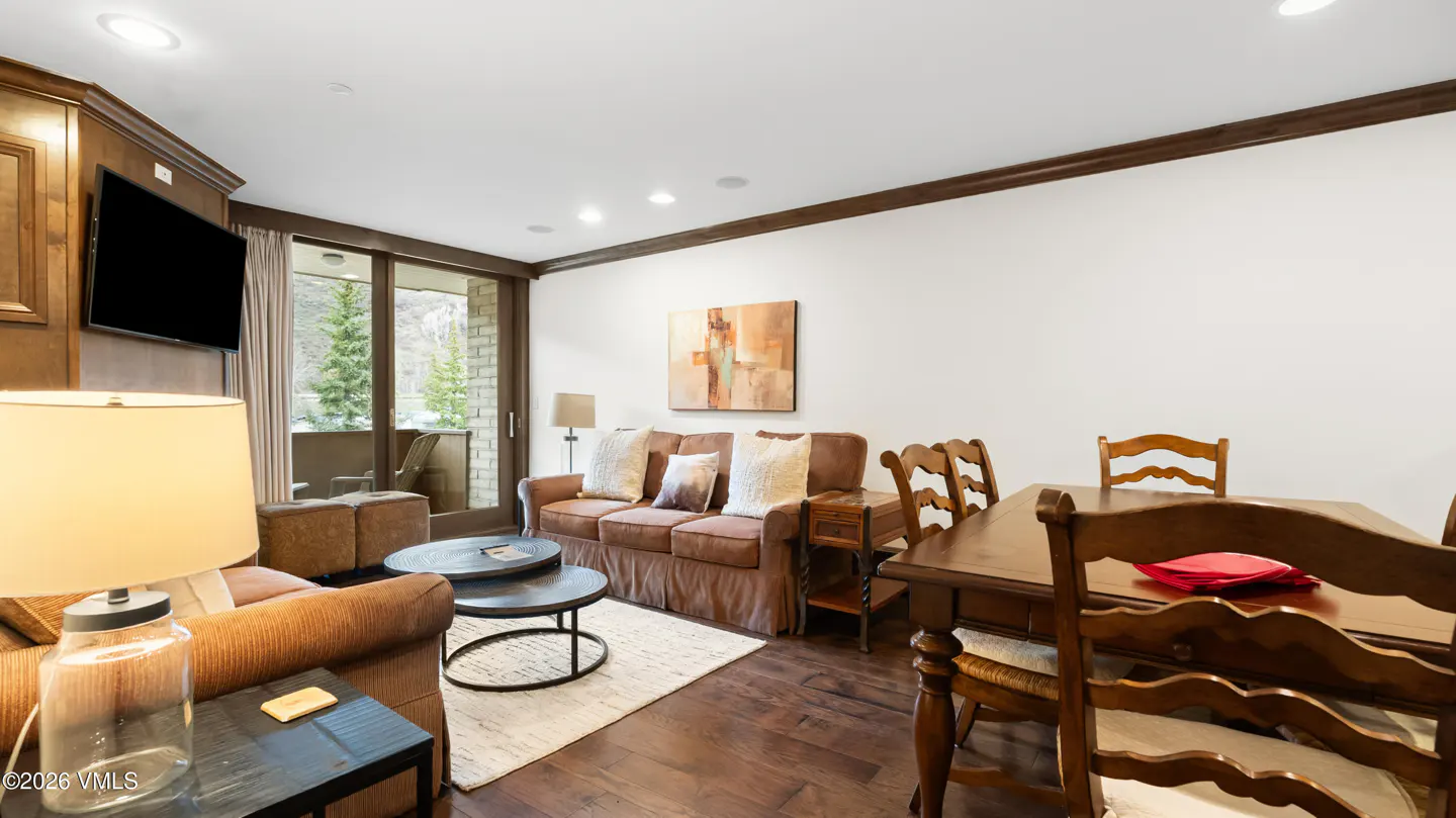 Living room with brown sofa, wood dining table, and balcony view of trees. The walls are white and the floors are dark wood.