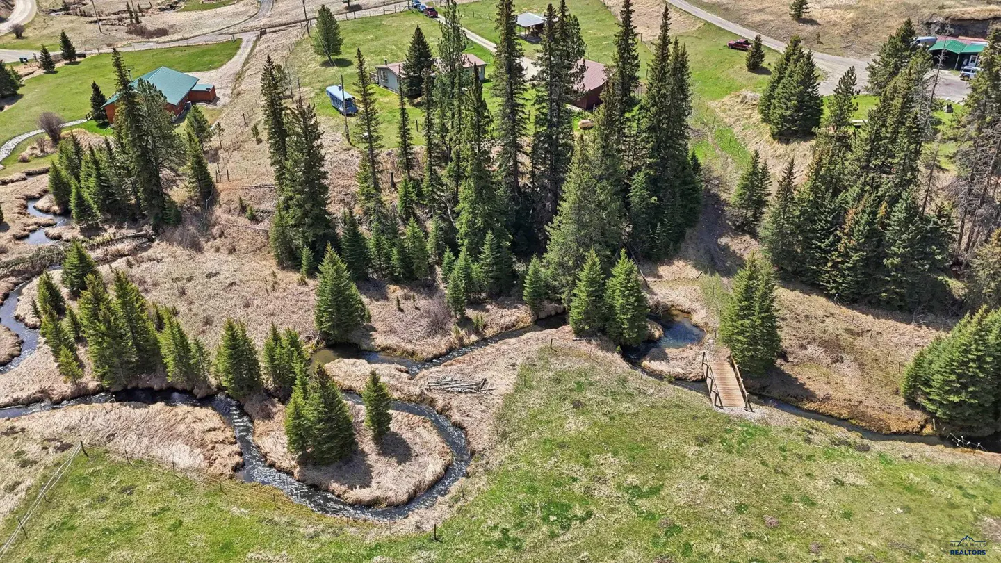 Aerial view of a winding creek, pine trees, and small cabins on a grassy hillside with a wooden footbridge.