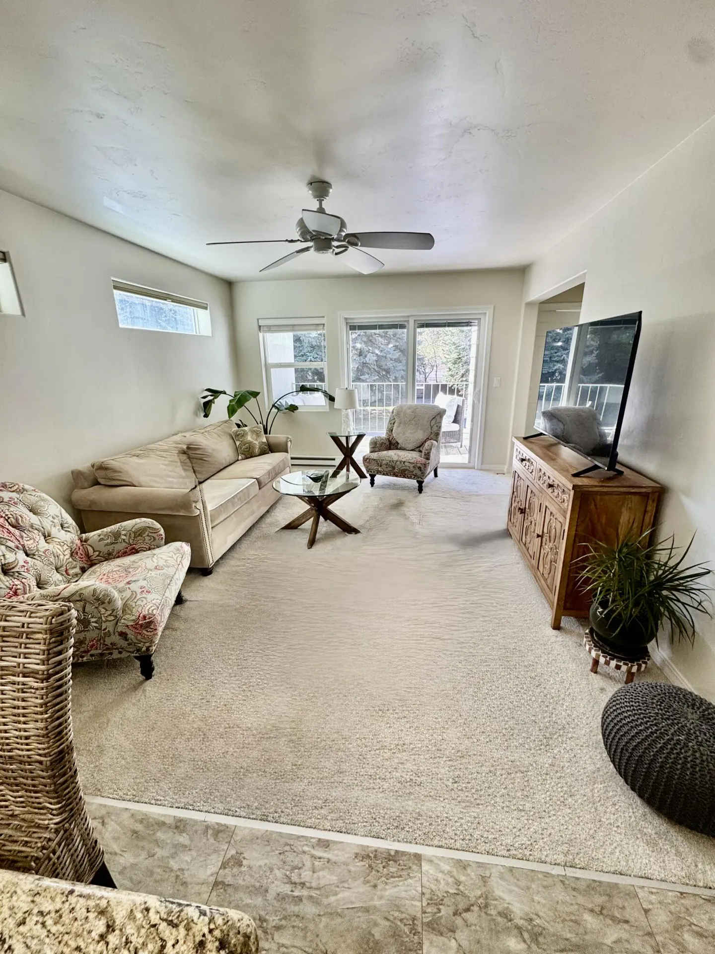 Living room with beige carpet, white walls, and a ceiling fan. A tan sofa, floral armchair, and glass coffee table furnish the space. A TV sits on a carved wooden cabinet.