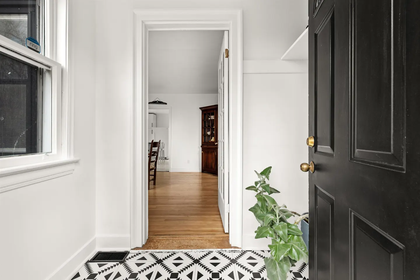 Open black front door with brass hardware, patterned tile floor, and view into a bright, white interior with wood floors.
