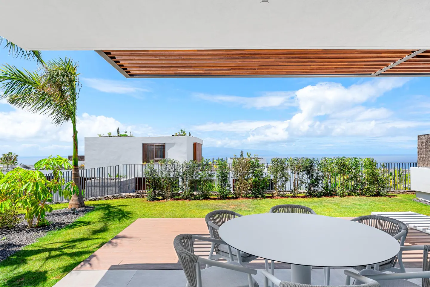 Outdoor patio with a white table and chairs, green lawn, and ocean view under a blue sky with clouds.
