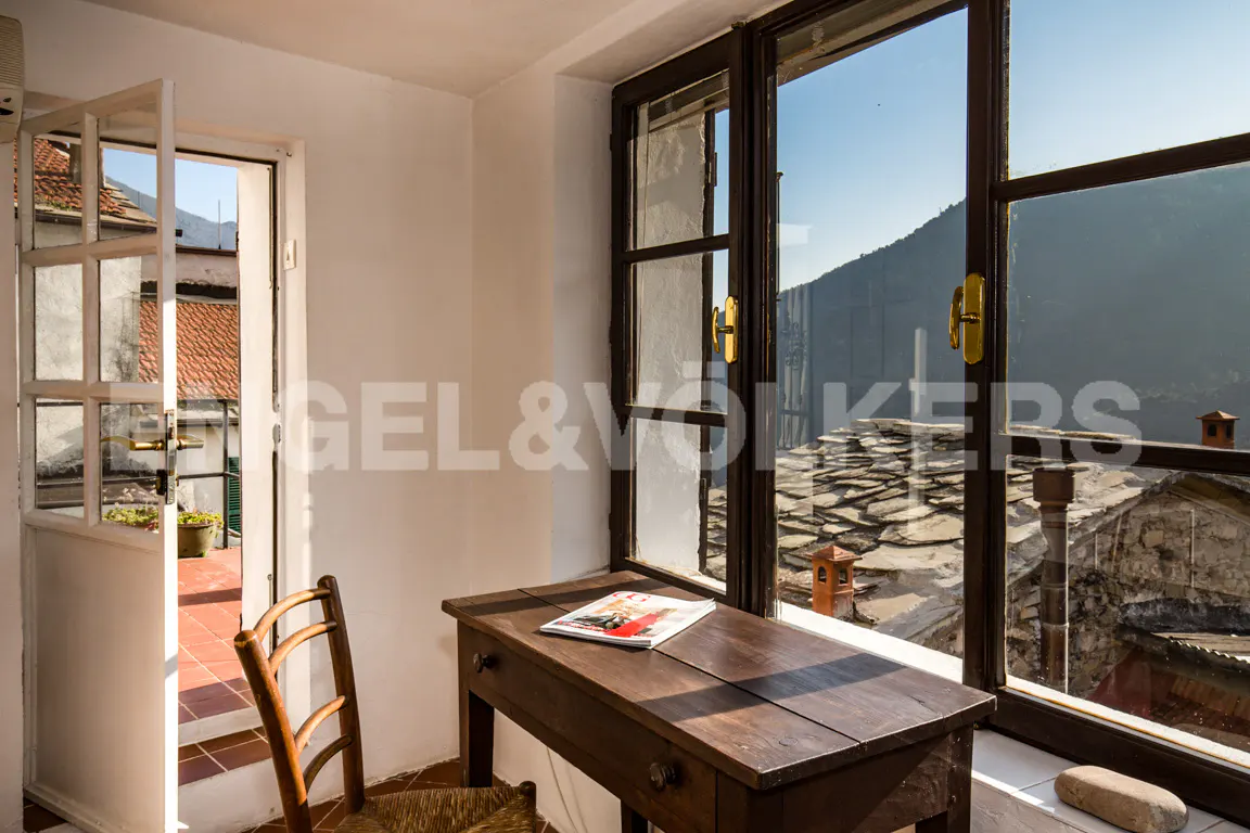 A bright room with a desk, chair, and open door. A magazine sits on the desk by a window with a view of rooftops and mountains.