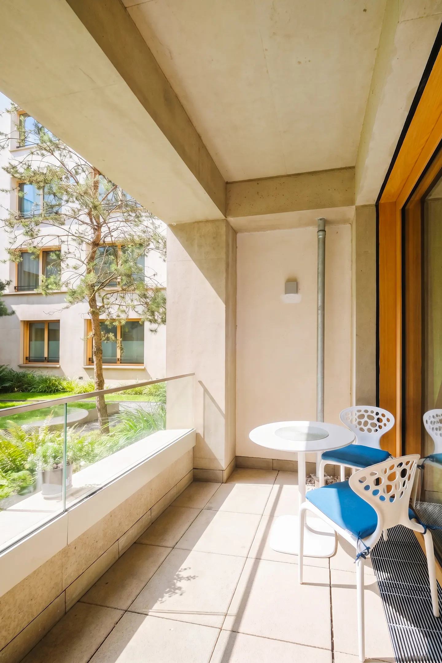 Sunlit balcony with white table and blue-cushioned chairs. Glass railing overlooks a green courtyard with a tree.