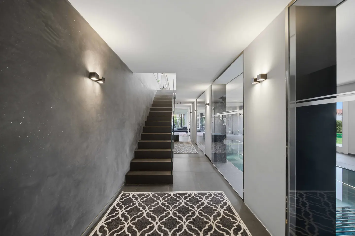 Modern hallway with gray walls, dark wood stairs, and a patterned rug. Mirrored panels line the right wall, reflecting light.