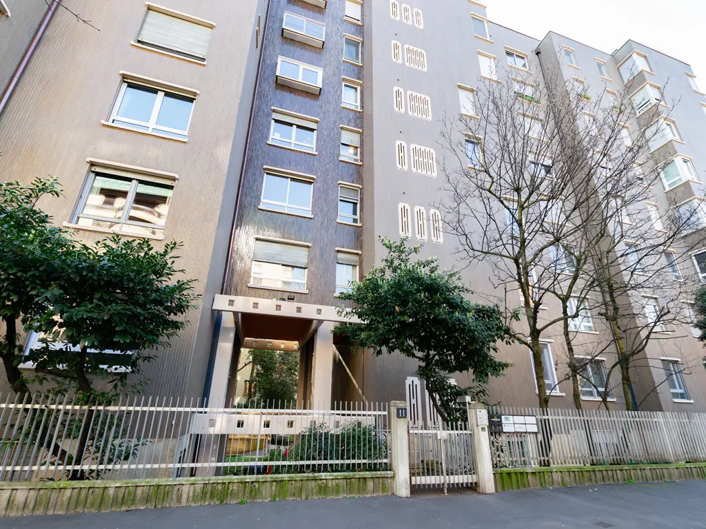 Exterior view of a tall, gray apartment building with a gated entrance and trees.