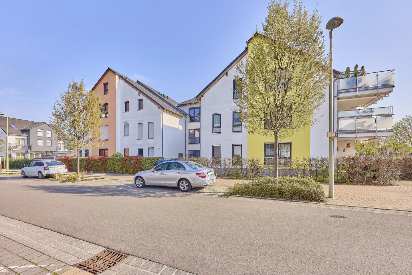 Exterior view of modern apartments with white and colored walls, balconies, parked cars, and trees under a blue sky.