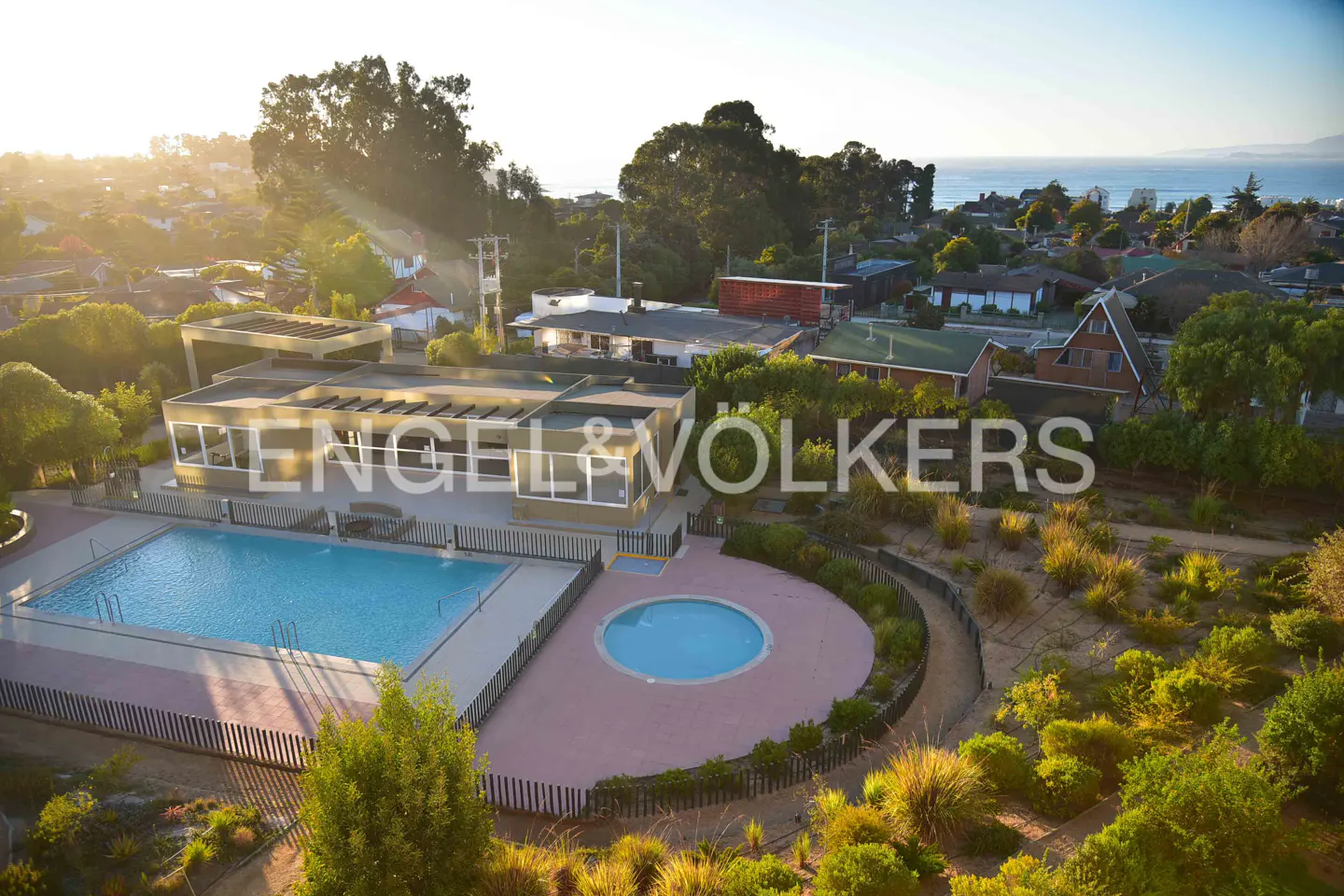 Aerial view of a property with a pool, patio, and landscaping, with houses and trees in the background. Ocean in the distance.
