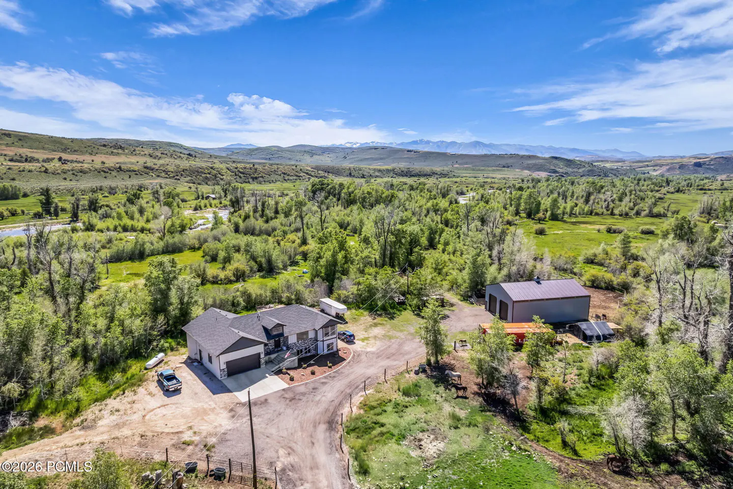 Aerial view of a rural home and barn set in a lush green valley with distant snow-capped mountains under a blue sky.