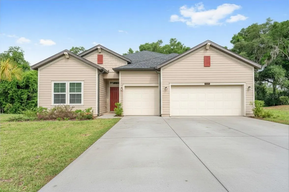 Beige single-story house with a gray roof, red door, and two-car garage. A concrete driveway leads to the house, with green grass on either side.