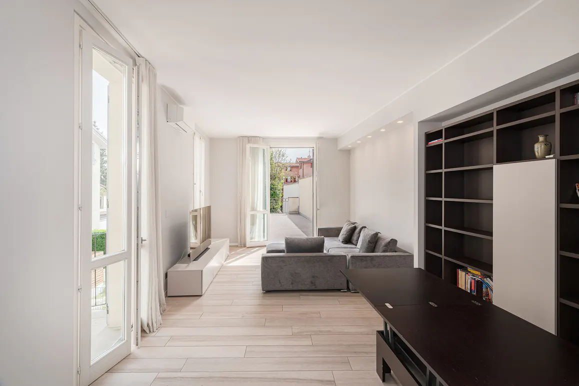 Bright living room with light wood floors, gray sofa, and dark wood bookcase. Balcony doors open to the outside.