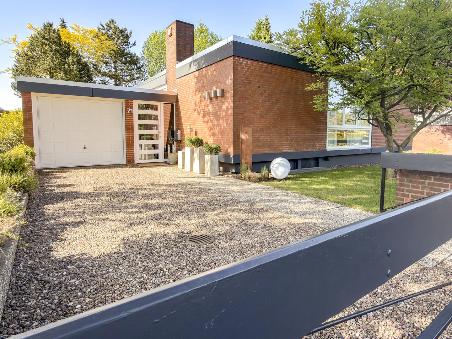 Mid-century modern red brick home with a white garage, gravel driveway, and green lawn under a clear blue sky.