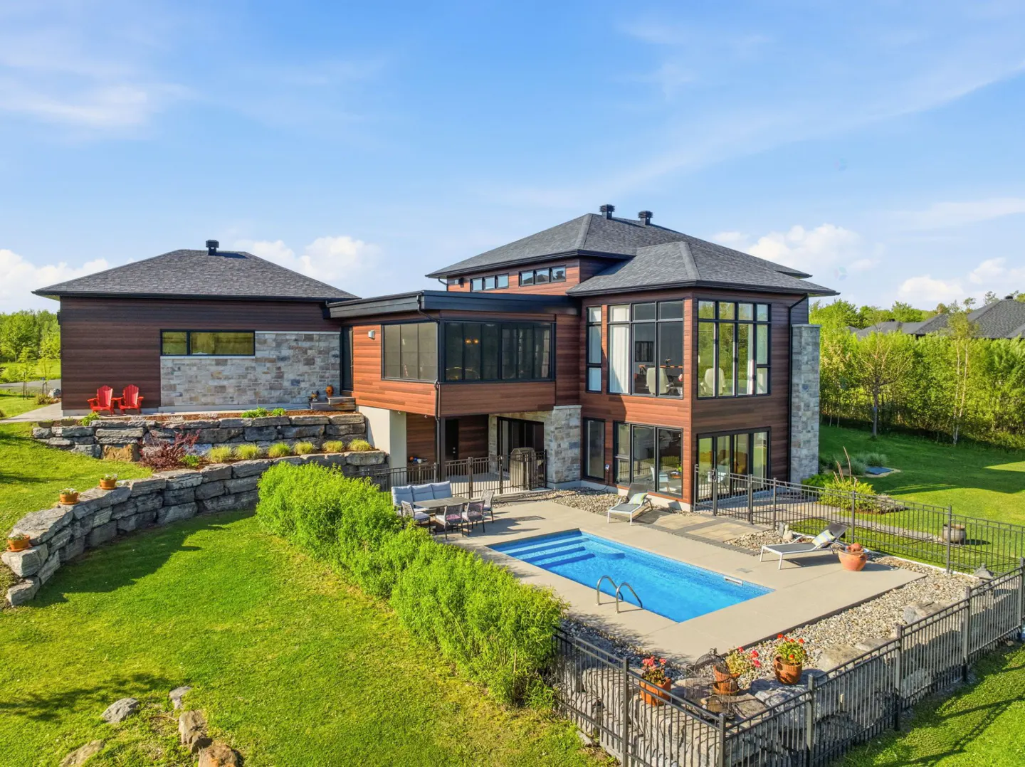 Modern home with wood and stone exterior, pool, patio furniture, and green lawn under a blue sky.