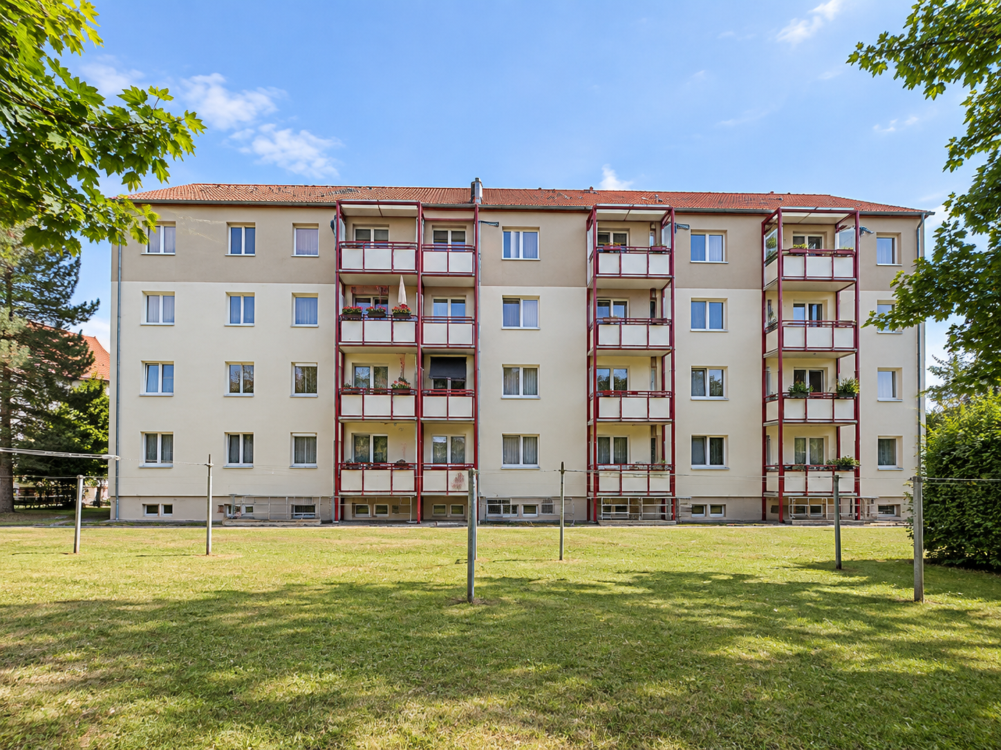 A four-story apartment building with beige walls, a red roof, and three vertical rows of balconies, set behind a grassy lawn. [AI-generated text]
