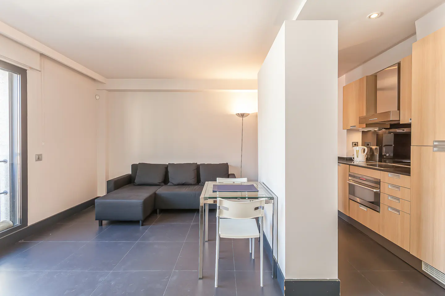 A modern apartment interior with a black sofa, glass table, and light wood kitchen cabinets.