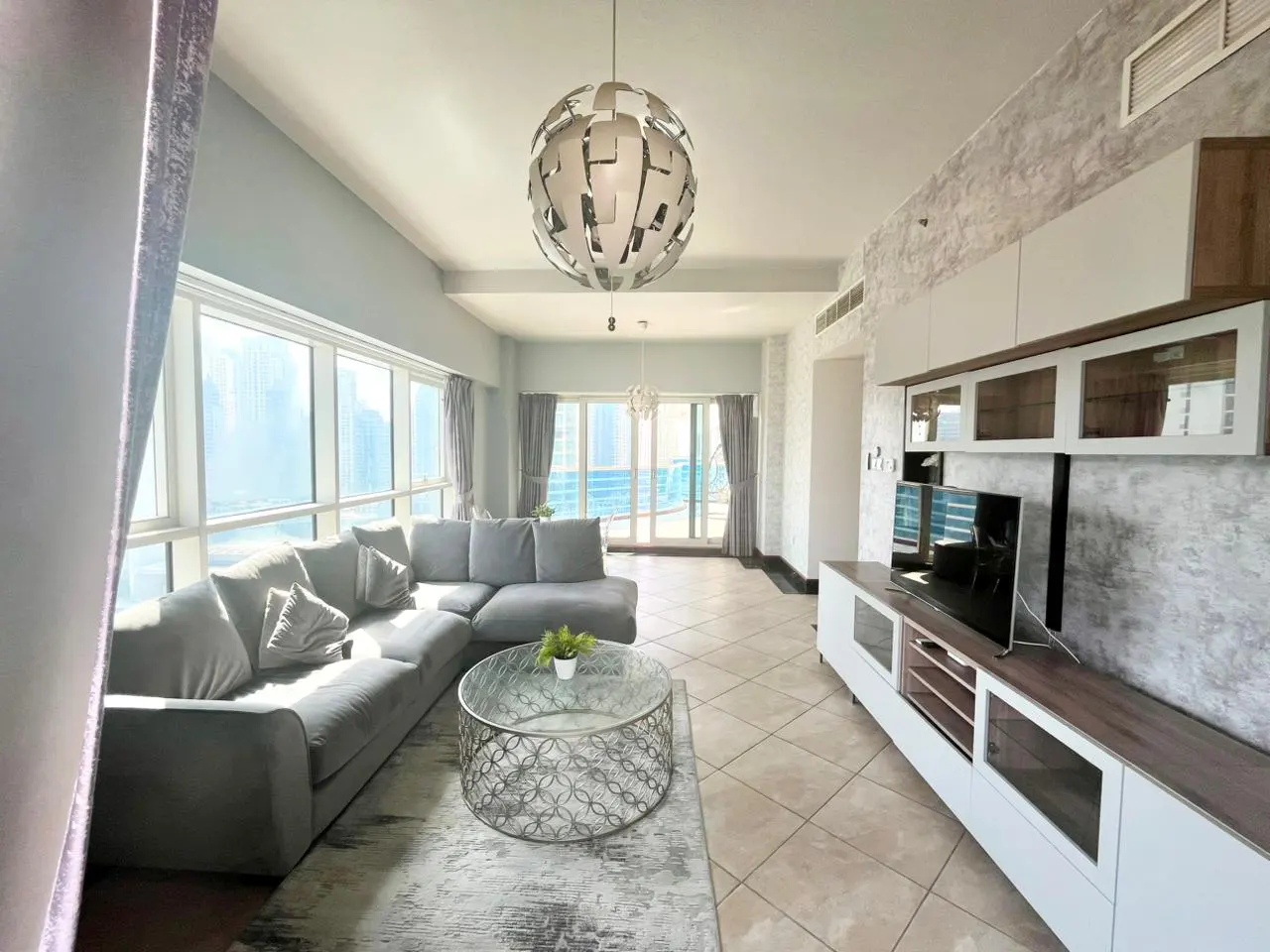 A modern living room with a gray sectional sofa, a glass coffee table, and a large window with city views. A spherical chandelier hangs from the ceiling.