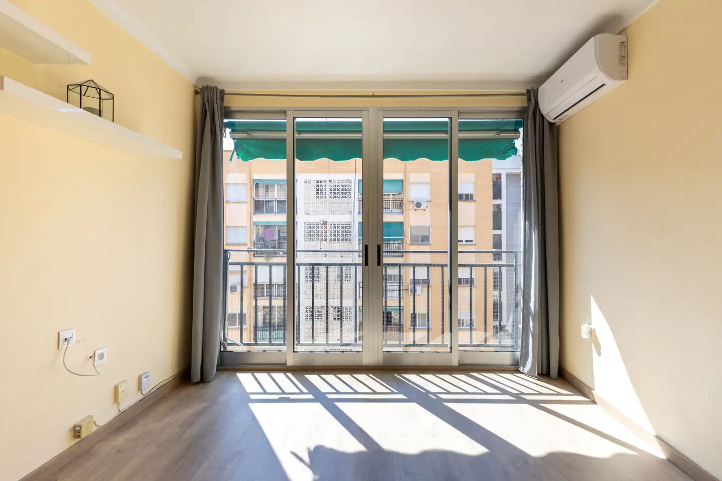Bright, empty room with yellow walls, wood floor, and sliding glass doors to a balcony. Gray curtains frame the doors.