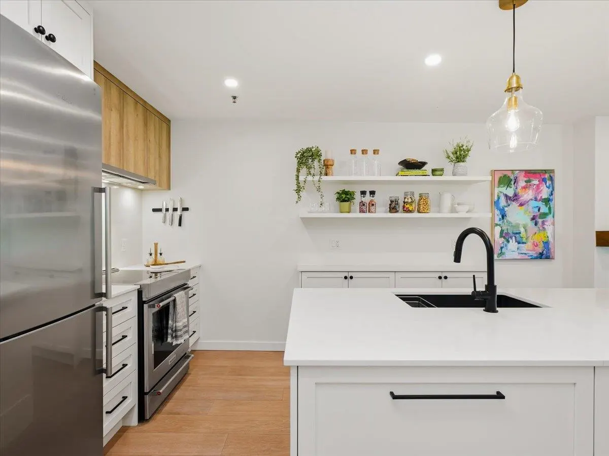 Bright kitchen with stainless steel fridge, white cabinets, and wood floors. A black faucet sits on a white island. Shelves display plants and jars.