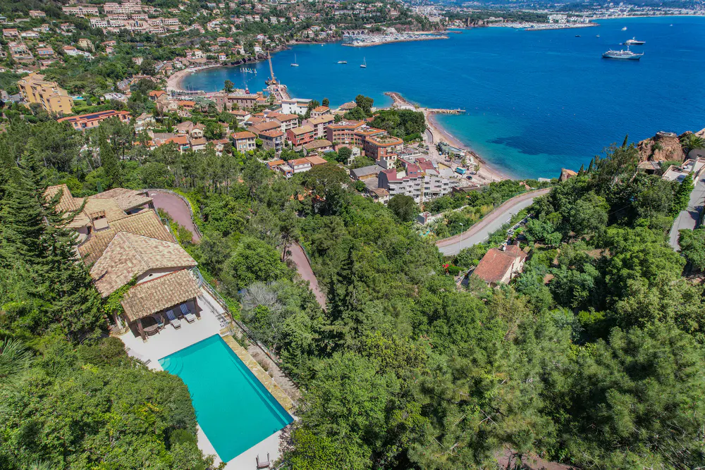 Aerial view of a villa with a pool overlooking a bay. The villa has a terracotta roof and is surrounded by trees. The bay has a beach and a town.