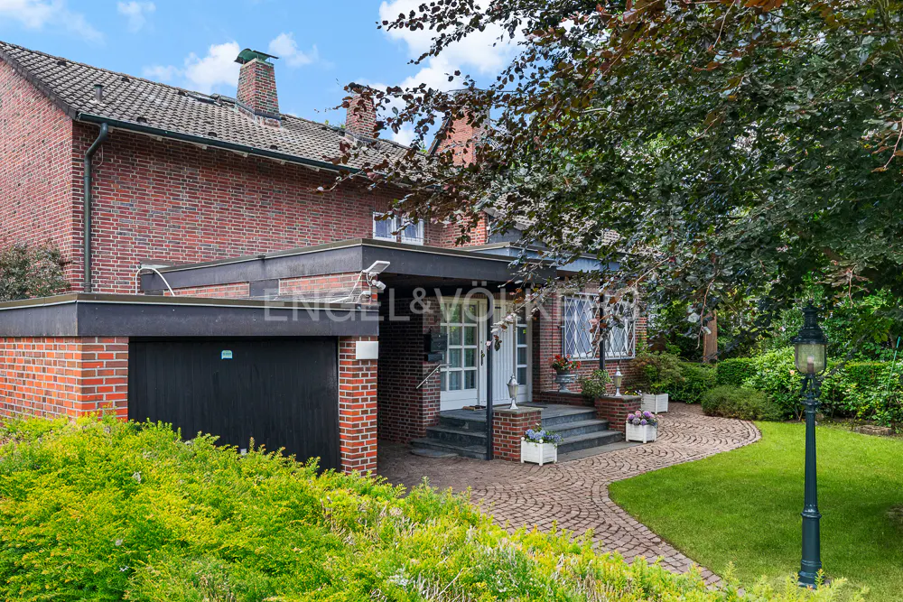 Exterior of a two-story brick house with a black garage door and a brick walkway leading to the front door. A green lawn and trees surround the house.