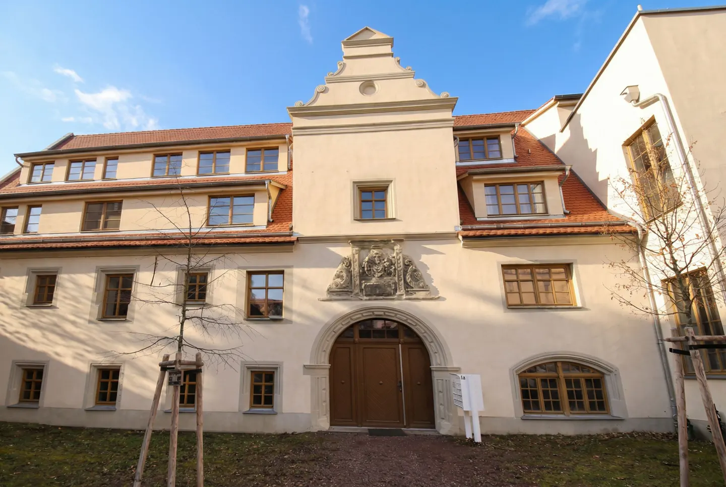 Exterior view of a cream-colored building with a red tile roof and brown arched doorway. An ornate stone carving sits above the entrance.
