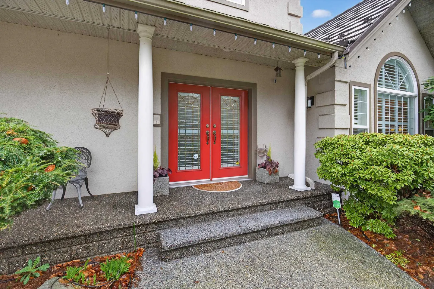 Front porch of a home featuring bright red double doors, white columns, a hanging planter, and lush green landscaping.